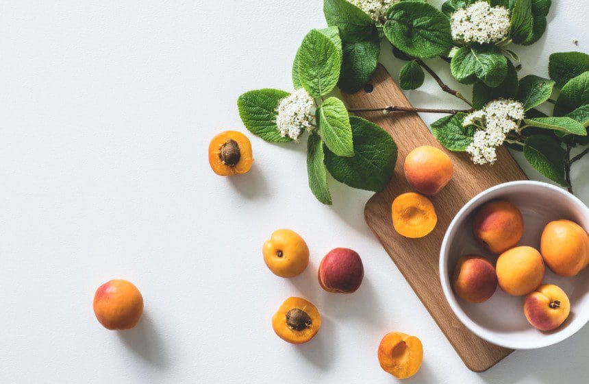 Donna Wright Designs Flat lay of fresh apricots and peaches on a wooden board beside clusters of white flowers, fragrant candles, and green leaves on a light background.