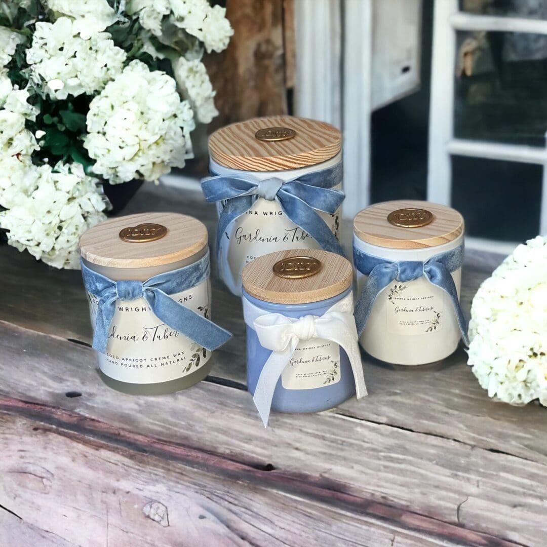 Donna Wright Designs Three elegant Gardenia & Tuberose scented candle jars with wooden lids, labeled "laura thomas co", displayed on a wooden surface, decorated with blue ribbons and accompanied by white hydrangeas.