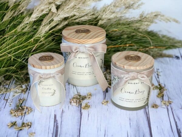 Three refreshing Ocean Breeze candles with wooden wicks by Donna Wright Designs in white glass vessels with beige velvet ribbon and wood lids sitting on a sandy wood table with sea oats in the background.