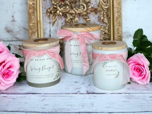 Three Spring Bouquet candles with wooden wicks by Donna Wright designs with frosted glass vessels, wooden lids and pink velvet ribbon on a white wood table with pink roses in the background.