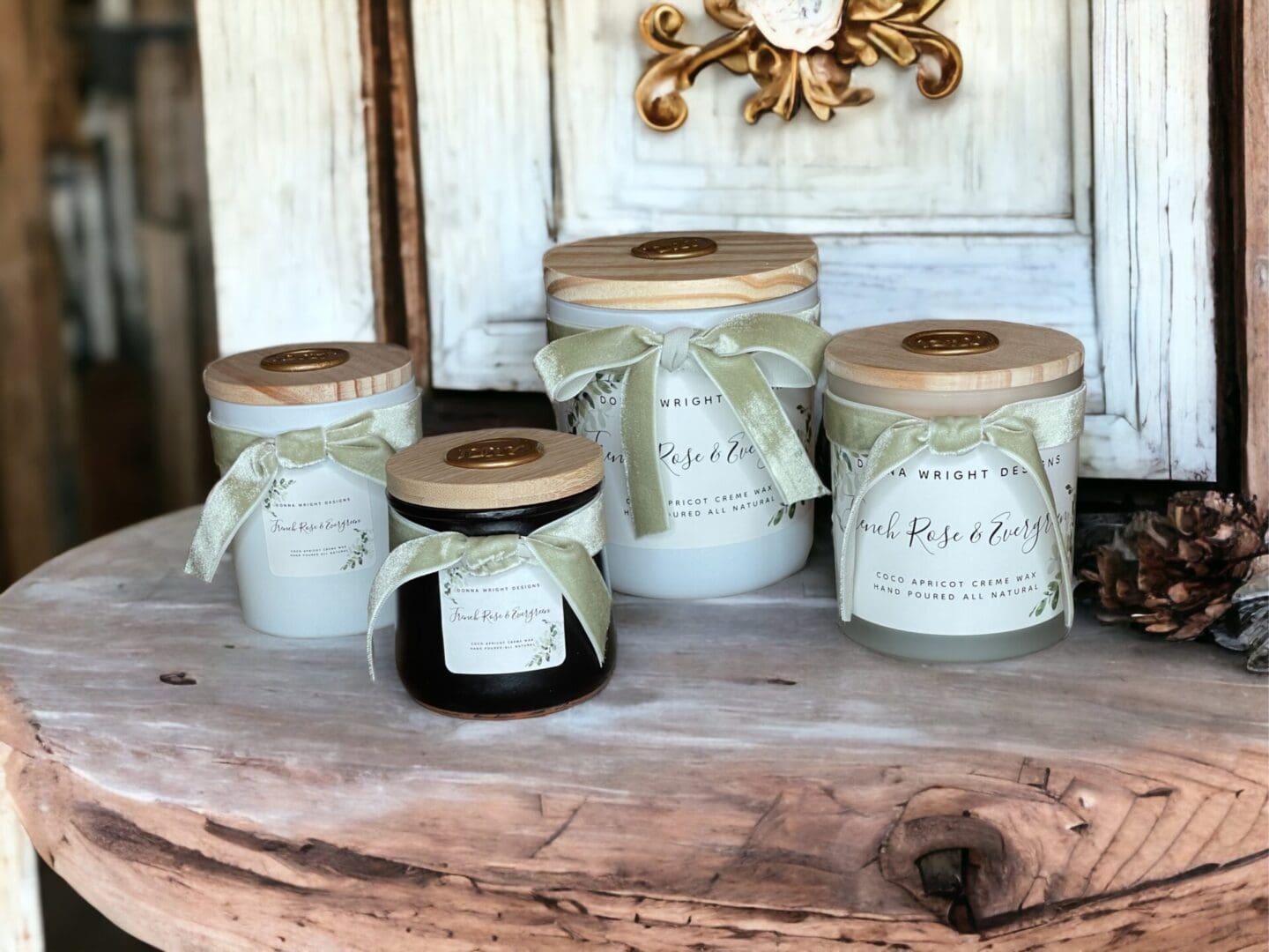 Donna Wright Designs Four scented French Rose & Evergreen candles with wooden lids on a rustic wooden table in front of a weathered white door. The candles are labeled with elegant script and feature a simple ribbon decoration.
