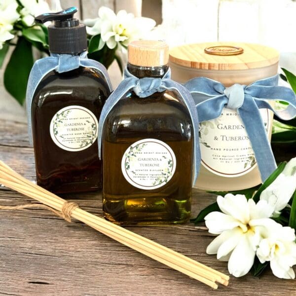 Two bottles with blue ribbon bows and a "Gardenia & Tuberose Candles" label are arranged alongside white gardenia flowers and reed sticks, creating a fresh, elegant display—ideal for anyone wanting the best gardenia scented candles.