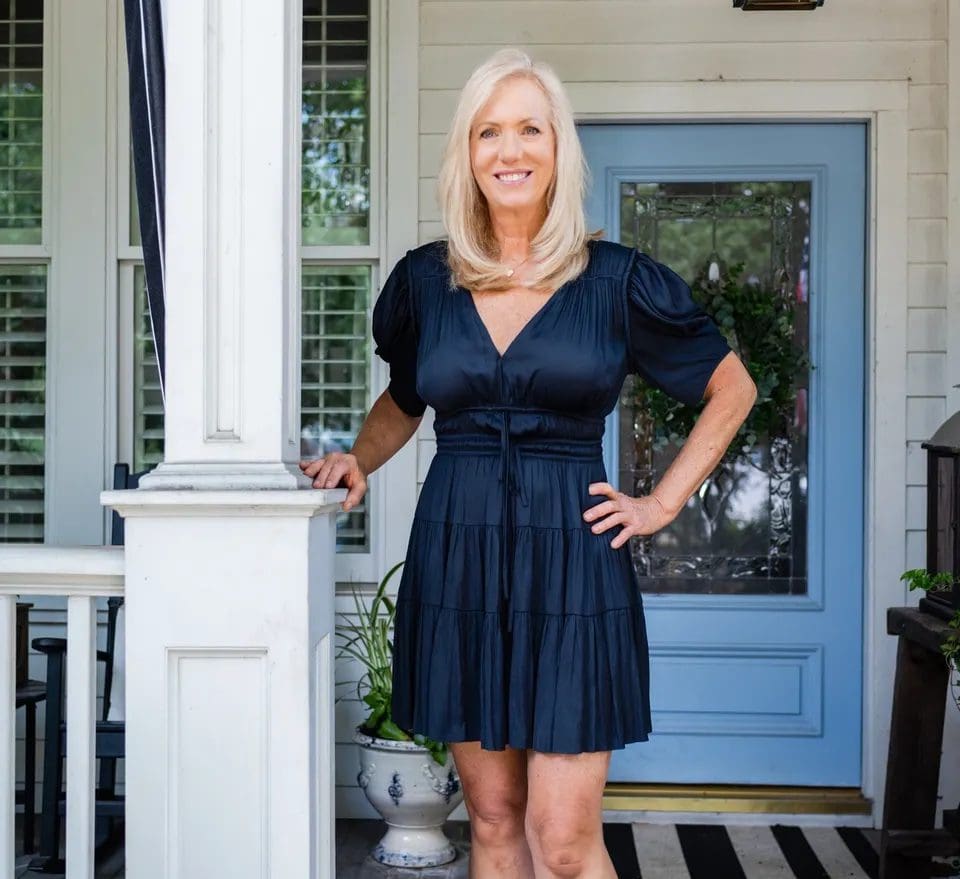 Donna Wright Designs A smiling woman with blonde hair, standing on a porch at the farmhouse, dressed in a navy blue dress, with her hand resting on a white pillar. The blue door and checkered doormat