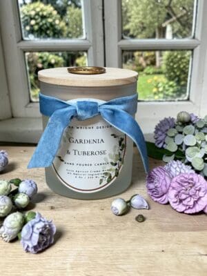 A frosted glass candle labeled "Gardenia & Tuberose" with a wooden lid and blue ribbon sits on a windowsill, surrounded by pastel purple and white artificial flowers, beneath a copy of header text. A garden is visible through the window.