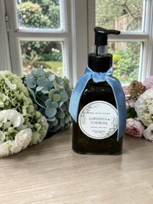 A bottle of gardenia and tuberose scented hand soap with a blue ribbon sits on a wooden table, surrounded by light pink and white flowers, like a copy of header notes from the garden beyond the window.