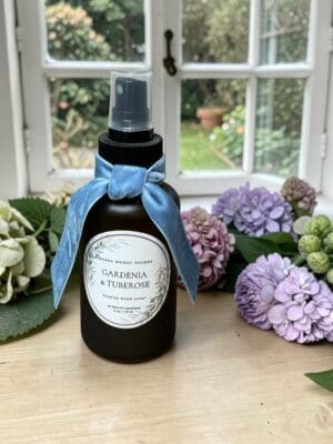 A black spray bottle labeled "Gardenia & Tuberose Scented Room Spray" with a blue ribbon sits on a windowsill, surrounded by pink, purple, and green hydrangeas beneath the soft header of an open window overlooking a garden.