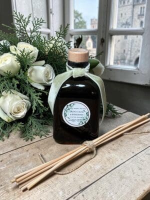 A glass bottle with a wooden cap and a green ribbon labeled “French Rose & Evergreen” sits on a rustic table with reed diffuser sticks, white roses, greenery, and a Copy of Header near a window overlooking a stone building.