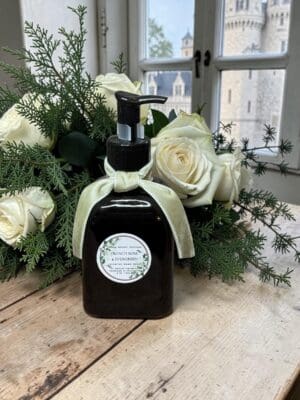 A dark pump bottle with a pale ribbon and floral label stands on a wooden surface near a window, the header of a stone castle visible outside, framed by white roses and greenery.