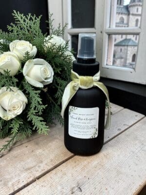 A black spray bottle with a light green ribbon sits on a wooden surface beside a header of white roses and greenery, in front of a window framing a blurred outdoor building.