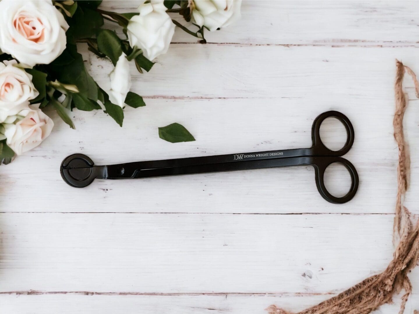 Donna Wright Designs A pair of black scissors lying next to twine and white roses on a wooden surface, accompanied by a Candle Wick Trimmer.