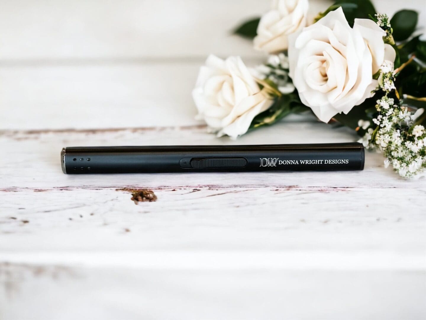 Donna Wright Designs A black stylus pen labeled "donna wright designs" lies on a white wooden surface next to a bouquet of white roses and a Candle Lighter - Refillable Butane.
