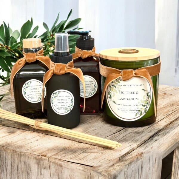 A header-worthy set of Fig Tree & Labdanum scented products, including a candle, spray, and diffuser with matching labels and brown velvet bows, elegantly displayed on a rustic wooden table amid lush greenery.