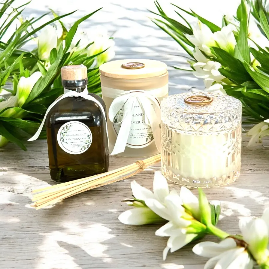 A glass jar candle with Woodland Lily & Vetiver Grass, a reed diffuser, and a bottle of oil are arranged on a white wooden surface, surrounded by white flowers and green leaves, creating a fresh, elegant spa-like setting—perfect for your home decor header.