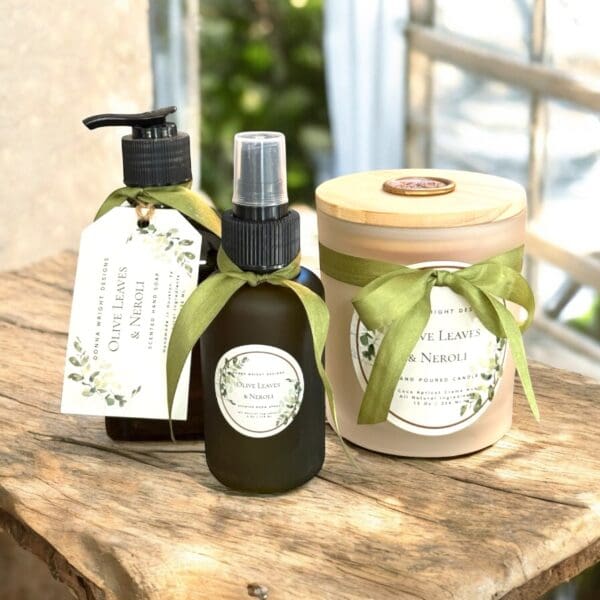 Header: Three Olive Leaves & Neroli scented products—a bottle with pump, a spray bottle, and a candle jar—decorated with green ribbons and botanical labels, sit on a rustic wooden surface near a sunlit window.