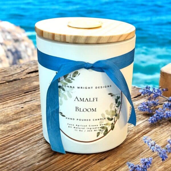 A white candle jar labeled "Amalfi Bloom" with a wooden lid and blue ribbon sits on a wooden surface by the sea, surrounded by lavender sprigs and shimmering blue water—a serene header for any coastal-inspired setting.