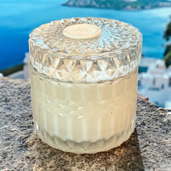 A decorative Amalfi Bloom candle in a glass jar with a textured, diamond-patterned surface sits on a stone ledge, serving as an elegant header to the scene filled with a white substance and set against a blurred coastal landscape and blue sea.