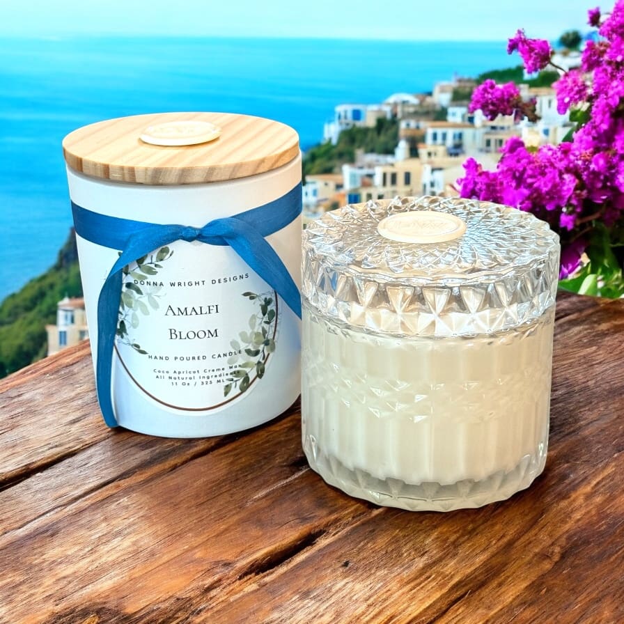 A decorative candle in a textured glass jar and its lidded container labeled "Amalfi Bloom" sit on a wooden table, beneath a vibrant header of pink flowers and a coastal village cascading down the hillside in the background.
