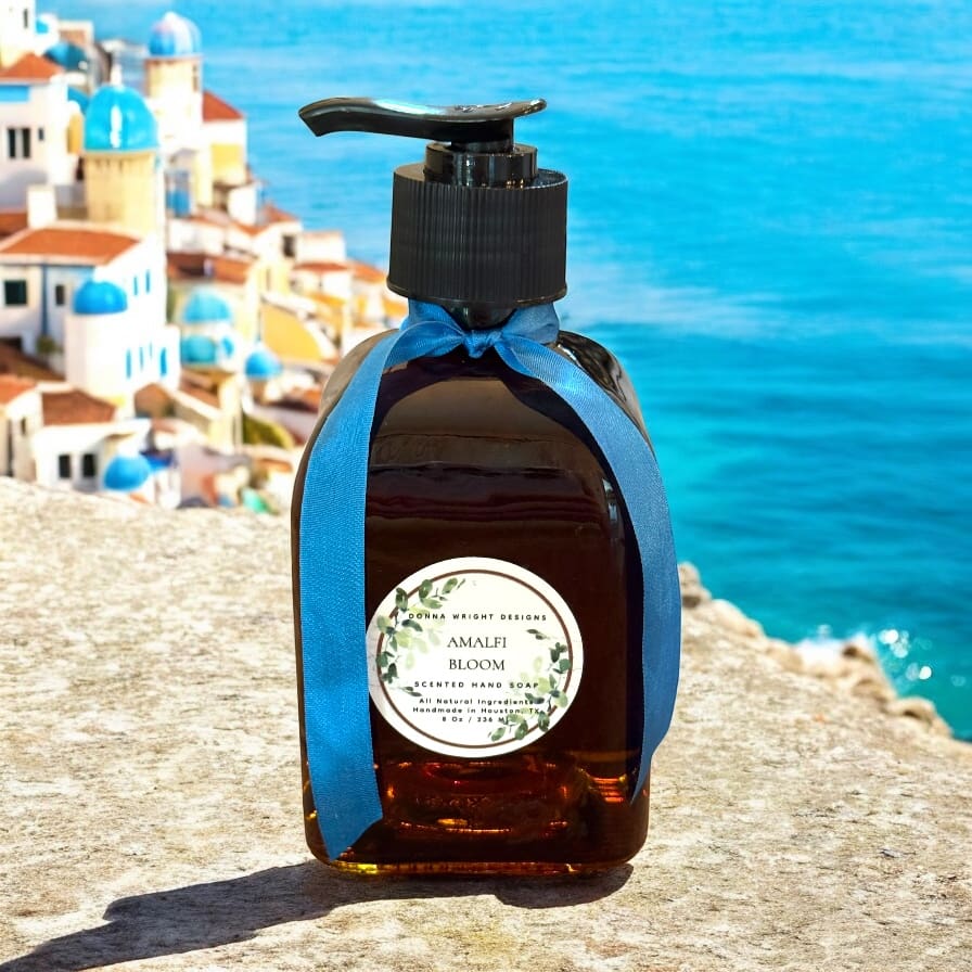 A brown soap dispenser with a blue ribbon stands on a stone ledge. A Header label reads “Malei Bloom,” set against white buildings with blue domes and the sea, evoking a Mediterranean coastal town.