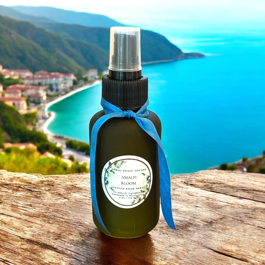 A dark spray bottle with a blue ribbon and a label reading "Amalfi Bloom" sits on a wooden surface, framed by a scenic coastal town, turquoise sea, and an elegant header highlighting its Mediterranean allure.