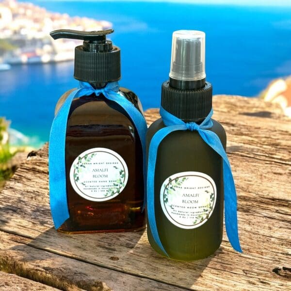 Two bottles labeled “Amalfi Bloom,” one with a pump and one with a spray, are tied with blue ribbons and set on rustic wood. In the blurred background header, bright blue coastal waters and charming buildings create a dreamy scene.