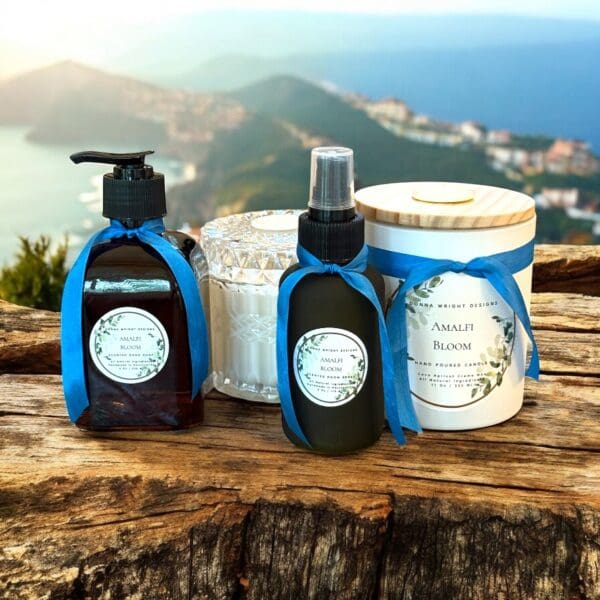 Skincare products and a candle labeled “Amalfi Bloom” sit on a rustic wooden surface with blue ribbons, set against a scenic backdrop of hills, water, and a coastal village—a serene Copy of Header moment.