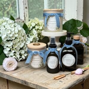 A charming header scene: “Gardenia & Tuberose” scented candles, sprays, and bottles adorned with blue ribbons rest on a rustic wooden table, complemented by white hydrangeas and lush greenery in the background.