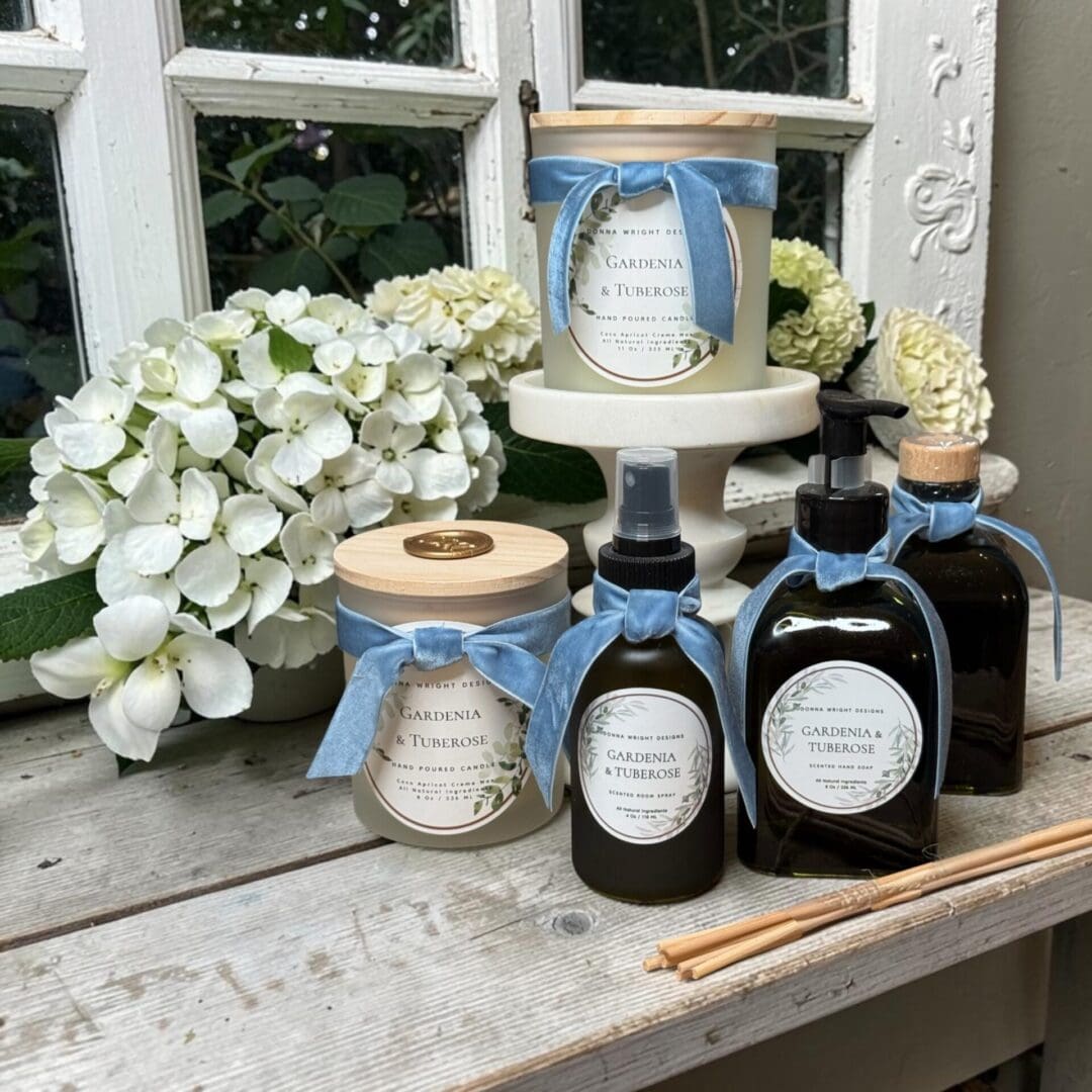 Header: A display of gardenia and tuberose scented candles, sprays, and diffusers in elegant jars with blue ribbons, set on a rustic wooden table with white hydrangeas and a vintage window in the background.