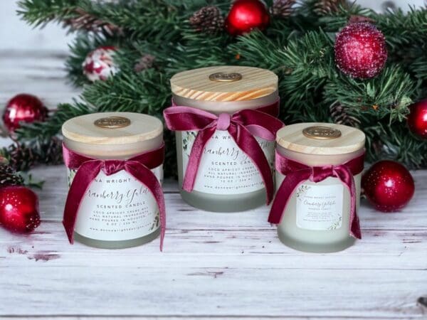 Three Cranberry Yuletide candles by Donna Wright Designs, luxury wooden wick candles in frosted glass vessels with pine wood lids & cranberry-colored velvet ribbon on a white wood table with red holiday ornaments and fresh pine branches in the background.