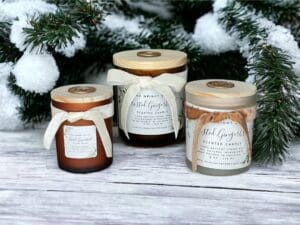 Three Frosted Gingerbread candles by Donna Wright Designs, luxury wooden wick candles in frosted amber vessels with ivory ribbon and wood lids on a white wood table with pine branches covered with snow in the background.