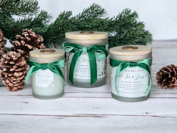 Three Noble Fir & Juniper Candles in frosted glass jars with wooden lids, each adorned with green ribbons, come in varying sizes and feature matching labels. These candles are displayed on a white wooden surface, surrounded by pinecones and evergreen branches.