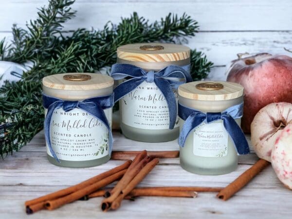 Three Warm Mulled Cider candles, luxury wooden wick candles with a blue bow on them sitting next to cinnamon sticks, apples & pine branches on a white wood table.