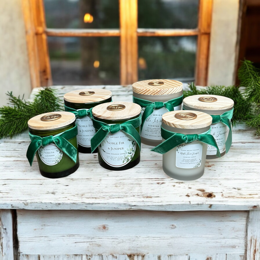 A rustic wooden table showcases six Noble Fir & Juniper Candles in glass jars, each topped with a wooden lid and adorned with a green ribbon. The candles come labeled with various scents, while decorative evergreen sprigs enhance the display. A window emitting a warm glow forms the backdrop.