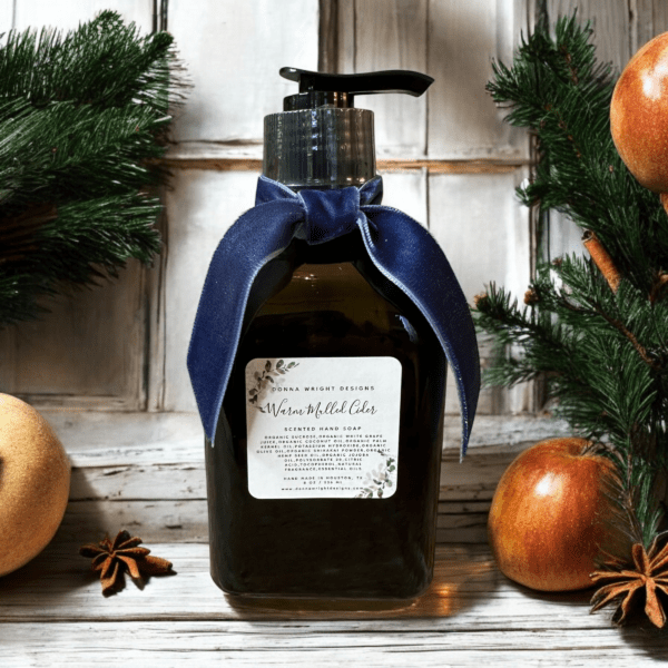 A bottle of Warm Mulled Cider Hand Soap by Donna Wright Designs sitting on a white table with apples, cinnamon and pine in the background next to a wooden window.