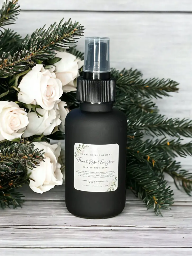 Donna Wright Designs A black spray bottle labeled "French Rose & Evergreen - Room Spray 4 Oz" surrounded by French roses and evergreen branches on a light wooden background.