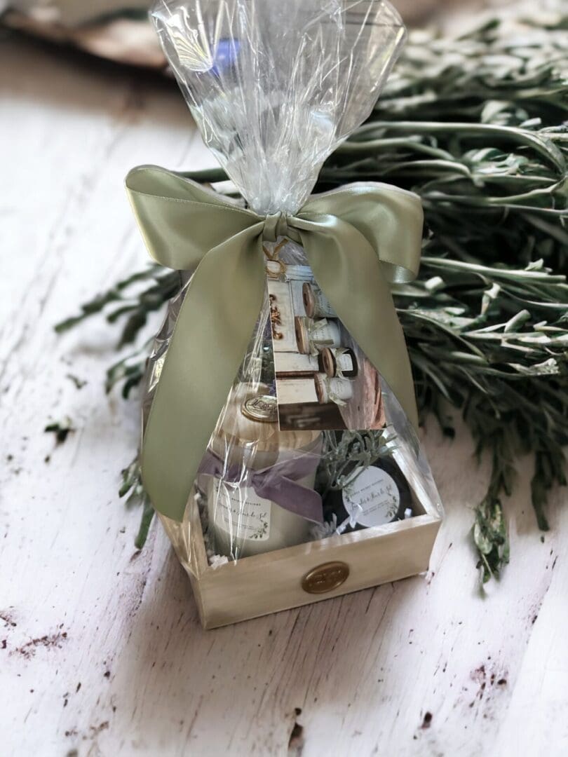 Donna Wright Designs A gift basket wrapped in clear cellophane with a green satin bow, containing various small items, placed on a wooden surface next to Noble Fir & Juniper - 4 Oz.