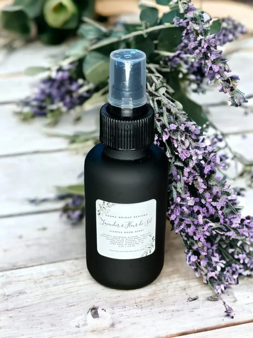 Donna Wright Designs A Noble Fir & Juniper - 4 Oz spray bottle with a white label sits in front of a sprig of purple flowers and juniper on a wooden surface.