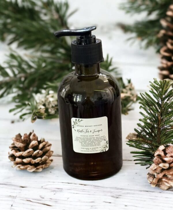 Noble Fir & Juniper hand soap by Donna Wright Designs is a luxury scented hand soap in an olive-colored bottle with black pump on a white wood table with pine cones & fir branches in the background