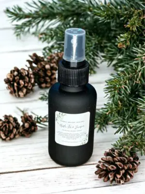 Noble Fir & Juniper room spray by Donna Wright Designs, the best pine scented room spray in an olive-colored glass bottle with black mist sprayer on a white wood table with pine cones & pine branches in the background.