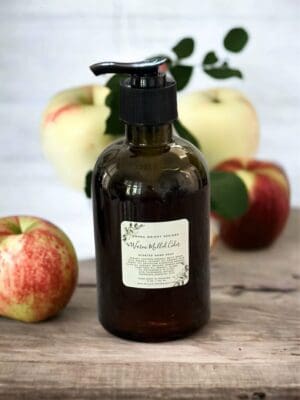 Warm Mulled Cider Hand. Soap by Donna Wright Designs, a luxury scented hand soap in an olive-colored bottle with black pump on a wooden desk with apples in the background.