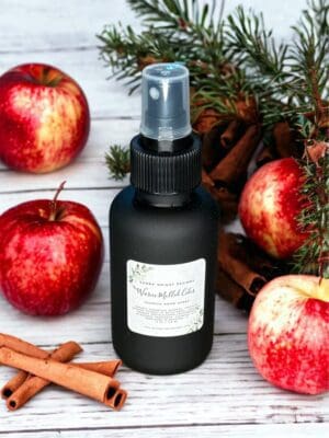 Warm Mulled Cider room spray by Donna Wright Designs, the best holiday room spray in a olive-colored glass bottle with black mist sprayer on a white wood table with apples, cinnamon & pine leaves in the background.