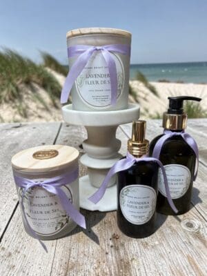 Lavender & Fleur de Sel Hand Soap, accented with purple ribbons, is displayed on a wood table near the beach, with sand and ocean in the background.