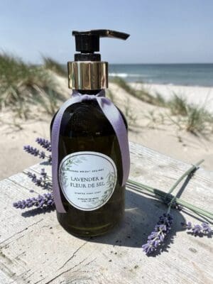 A bottle of Lavender & Fleur de Sel Hand Soap, adorned with a purple ribbon, rests on a wooden surface by the beach, surrounded by fragrant lavender sprigs and set against a backdrop of sand dunes and ocean waves.