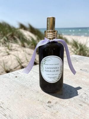 A dark glass spray bottle featuring a gold nozzle and accented with a lavender ribbon rests on a wooden surface near sandy beach grass, with the ocean and blue sky beyond. The label displays “Lavender & Fleur de Sel Hand Soap,” capturing the luxurious essence of this elegant coastal-inspired product.
