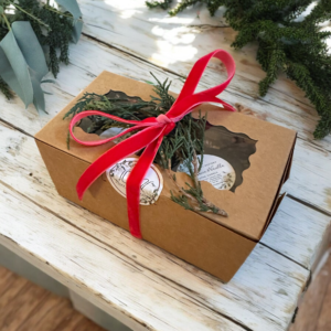 The Seasonal Small gift set, featuring two seasonal travel candles & a decorative holiday set of matches in a see-through window and tied with a red velvet ribbon, is adorned with seasonal greenery. It rests on a rustic white wooden table surrounded by green foliage, creating a festive and natural presentation.