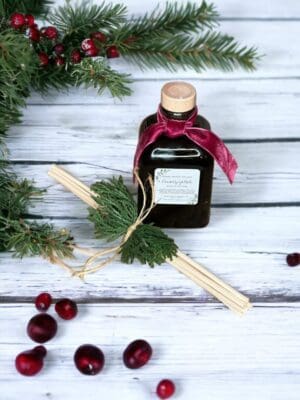 Cranberry Yuletide reed diffuser by Donna Wright Designs, the best Cranberry scented reed diffuser in an olive-colored glass bottle with cork lid sitting on a white wood table with fresh cranberries & evergreen in the background.