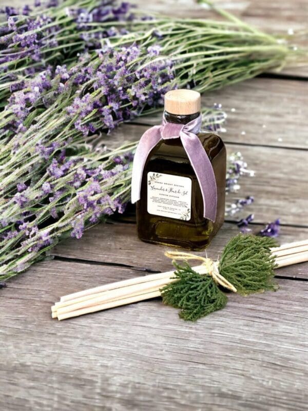 Lavender & Fleur de Sel reed diffuser 6 oz by Donna Wright Designs presented in an olive-colored glass bottle with cork lid, rattan reed sticks & purple ribbon on wood table with fresh lavender in the background.