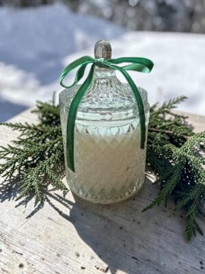 The Noble Fir & Juniper Reed Diffuser, housed in a decorative glass jar with a lid and green ribbon, rests on a wooden surface adorned with evergreen branches under sunlight, while snow softly blurs the background.