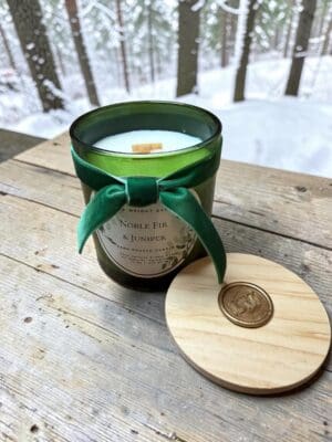 A green glass vessel labeled “Noble Fir & Juniper Reed Diffuser” with a green velvet bow sits on a rustic wooden surface, echoing the invigorating ambiance of a luxury fir fragrance. Its wooden lid, adorned with a wax seal, rests nearby, while snowy trees create a blurred backdrop that enhances the diffuser’s elegant, wintry charm.
