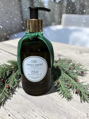A bottle of hand wash adorned with a green ribbon sits on a wooden table amid evergreen sprigs and red berries, with snowflakes softly falling in the background—the perfect pairing to the Noble Fir & Juniper Reed Diffuser for an elegant touch of winter luxury.