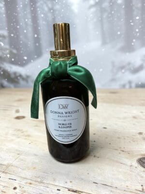 A dark glass spray bottle with a gold top and green ribbon, labeled “Donna Wright Designs Noble Fir & Juniper Home Fragrance Spray,” sits on a wooden surface against a snowy forest backdrop, mirroring the refined design of the Noble Fir & Juniper Reed Diffuser—an inviting complement to any luxury home fragrance collection.